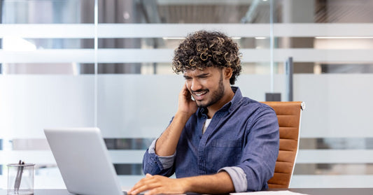 A curly-haired man in a denim shirt sitting at a desk using a laptop, holding his right ear in discomfort.