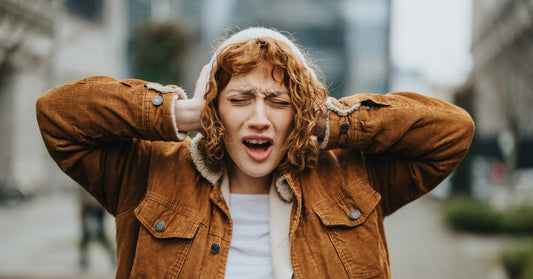 A woman wearing a brown jacket stands outside and presses her hands over her ears. Her expression is distressed.