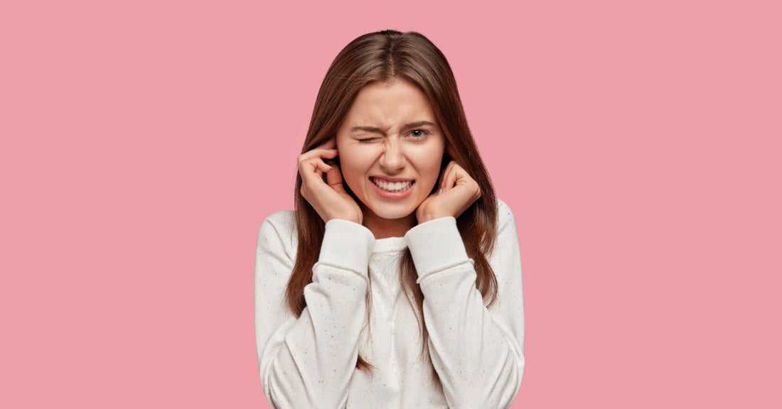 A woman wearing a white shirt stands against a pink background. She's putting her fingers in her ears and grimacing.