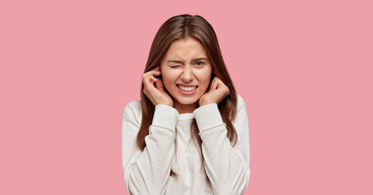 A woman wearing a white shirt stands against a pink background. She's putting her fingers in her ears and grimacing.