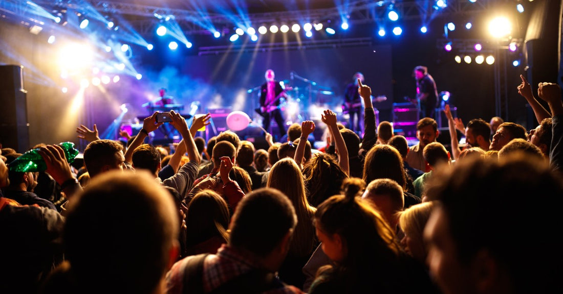 A concert stage glows with purple and blue lights in front of a crowd of people putting their arms up.