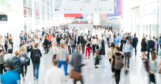 A large crowd of people walks through a bright convention center lobby beneath overhead directional signs.