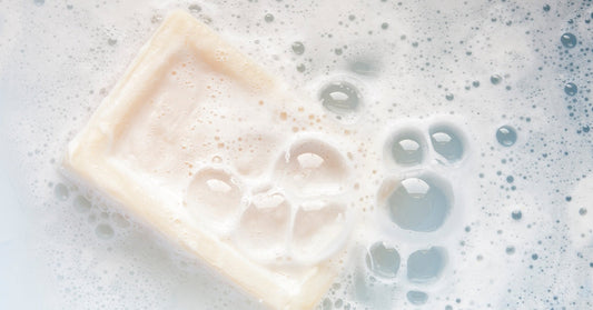 A bar of soap sits partially submerged in foamy water, surrounded by bubbles and soft white suds.